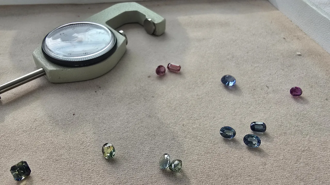 Loose faceted sapphires in blue, pink, purple and green scattered on a beige tray beside a dial thickness gauge